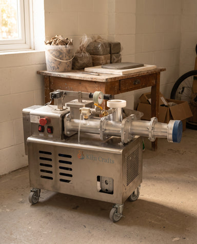 Kiln Crafts Mini De-Airing Pugmill set up at home in a garage, with a bucket of clay scraps, bagged clay and a plaster drying slab on a wooden workbench behind it