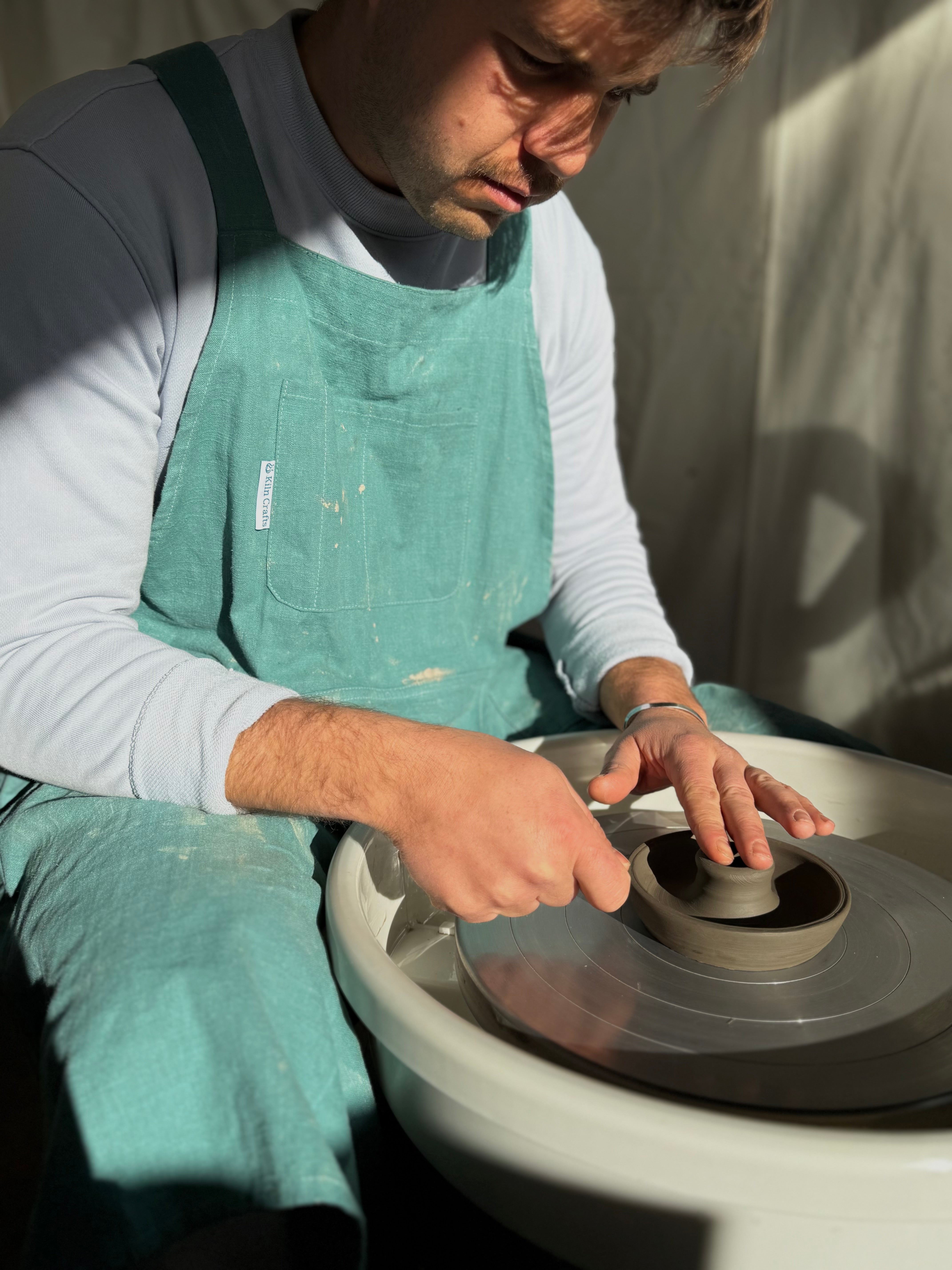 A potter in a green apron carefully shapes a clay bowl on a spinning pottery wheel in a sunlit studio