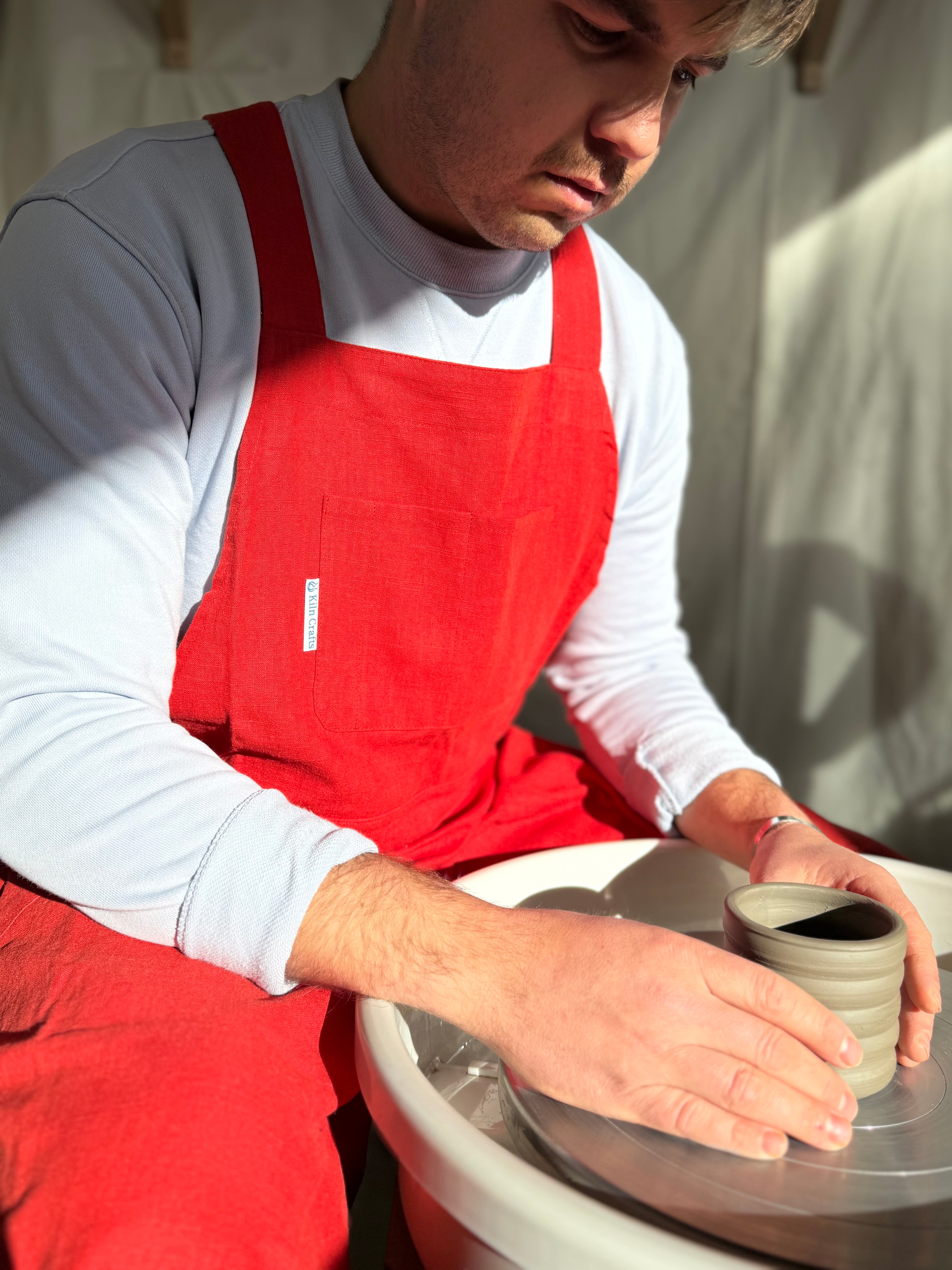 A potter in a red apron holds a ribbed clay vessel over a white pottery wheel in a sunlit studio.