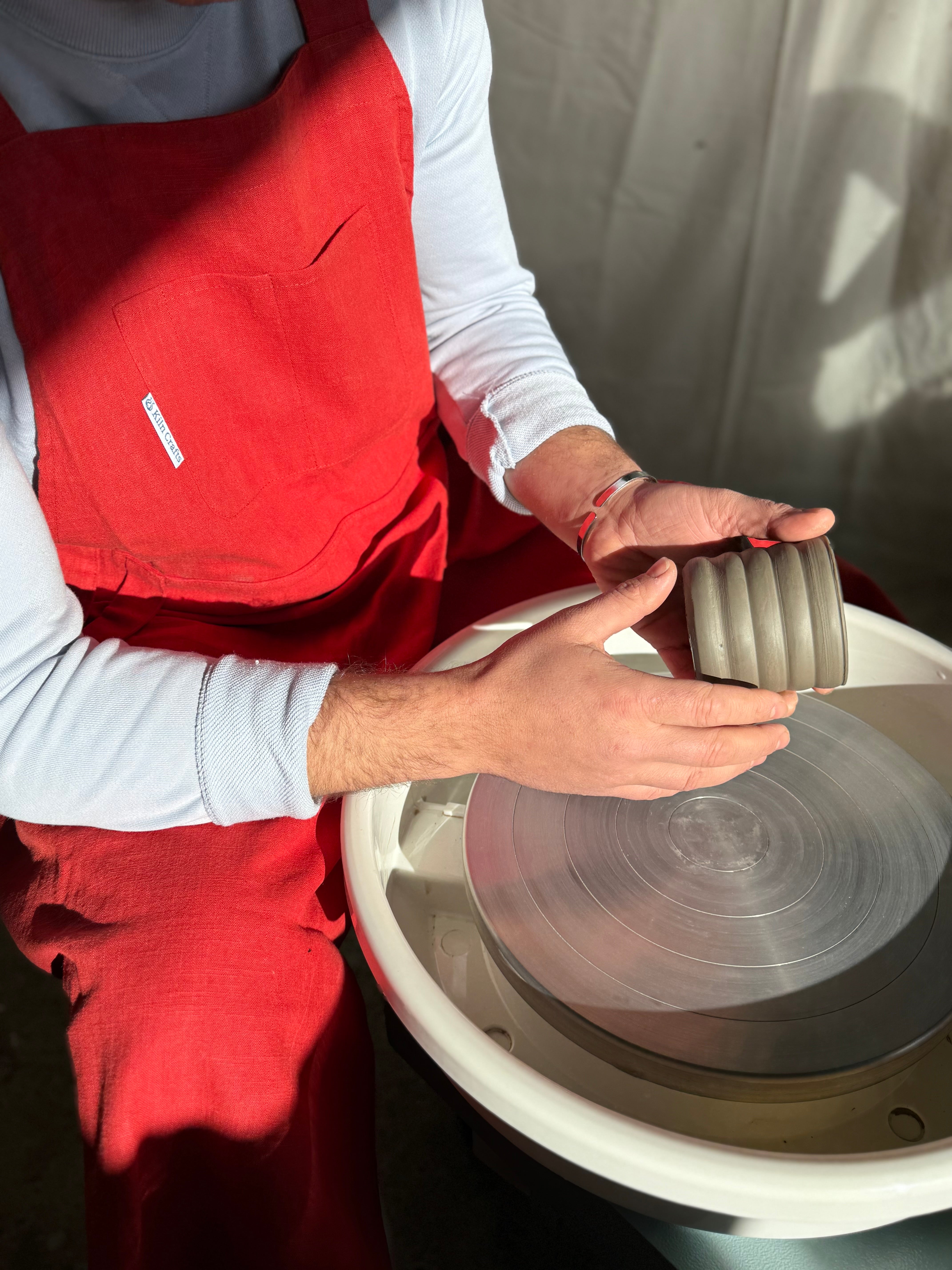 A potter in a red apron holds a ribbed clay vessel over a white pottery wheel in a sunlit studio.