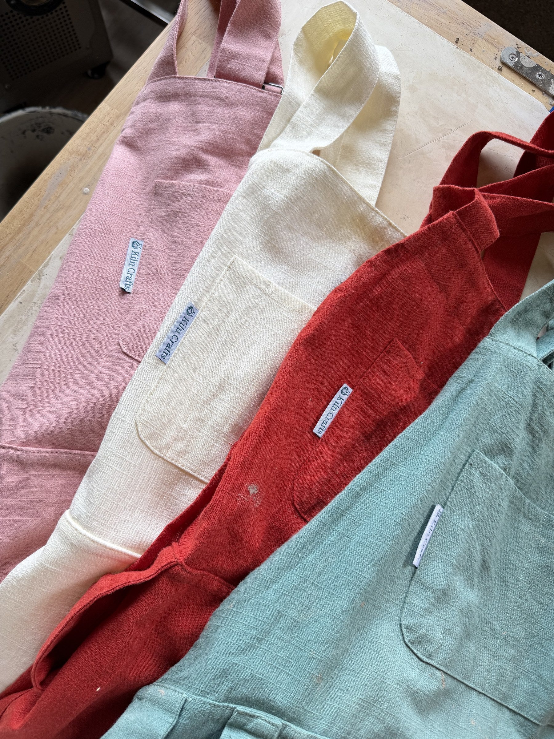 Kiln Crafts Craft Aprons in all four colours - Pink, Milk, Red and Teal - laid out together on a workbench