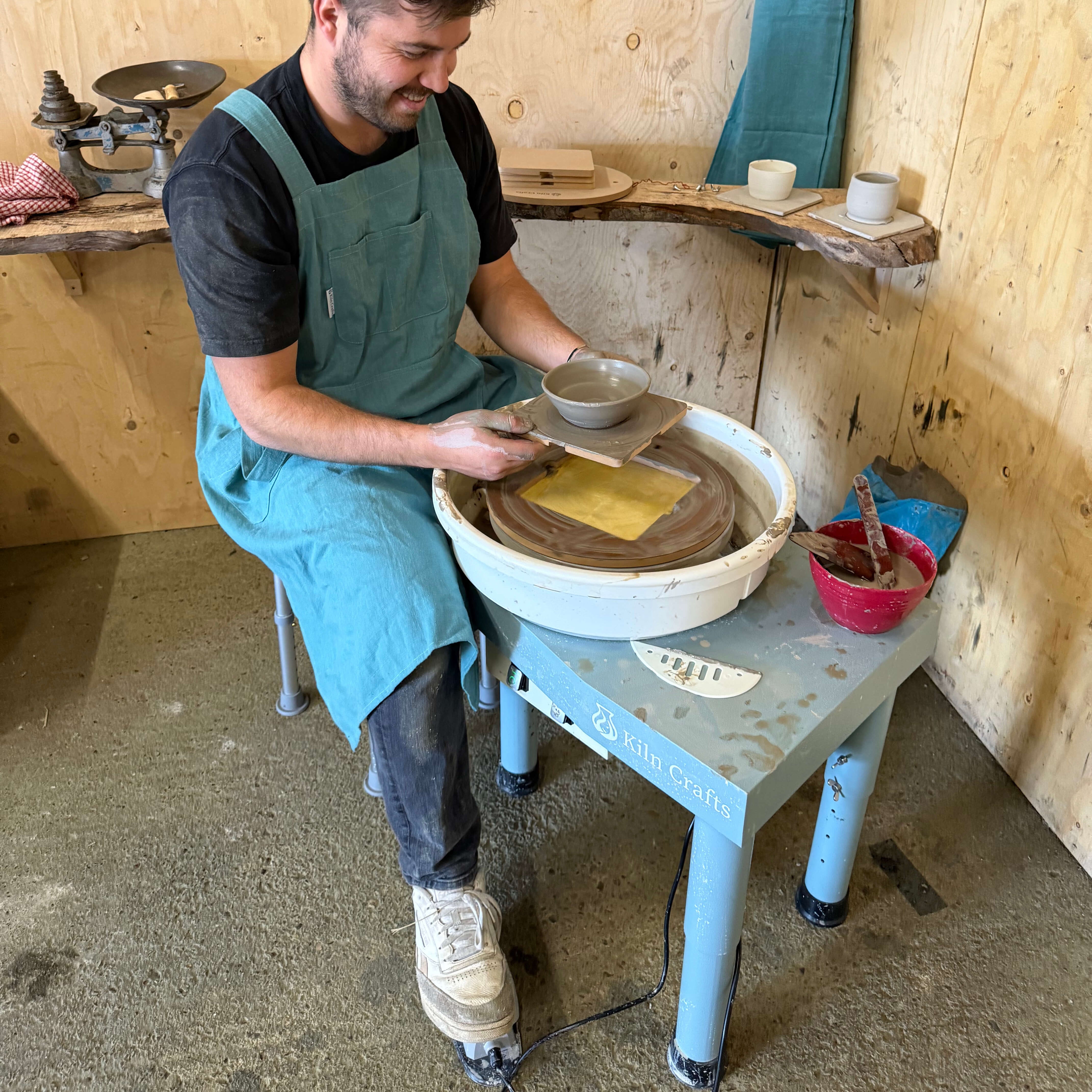 A male potter wearing a light blue apron sits at a Kiln Crafts pottery wheel in a rustic wood-panelled studio. He is smiling while holding a freshly thrown clay bowl on a wooden bat, with finished ceramic pieces and traditional scales visible on wooden shelves in the background.