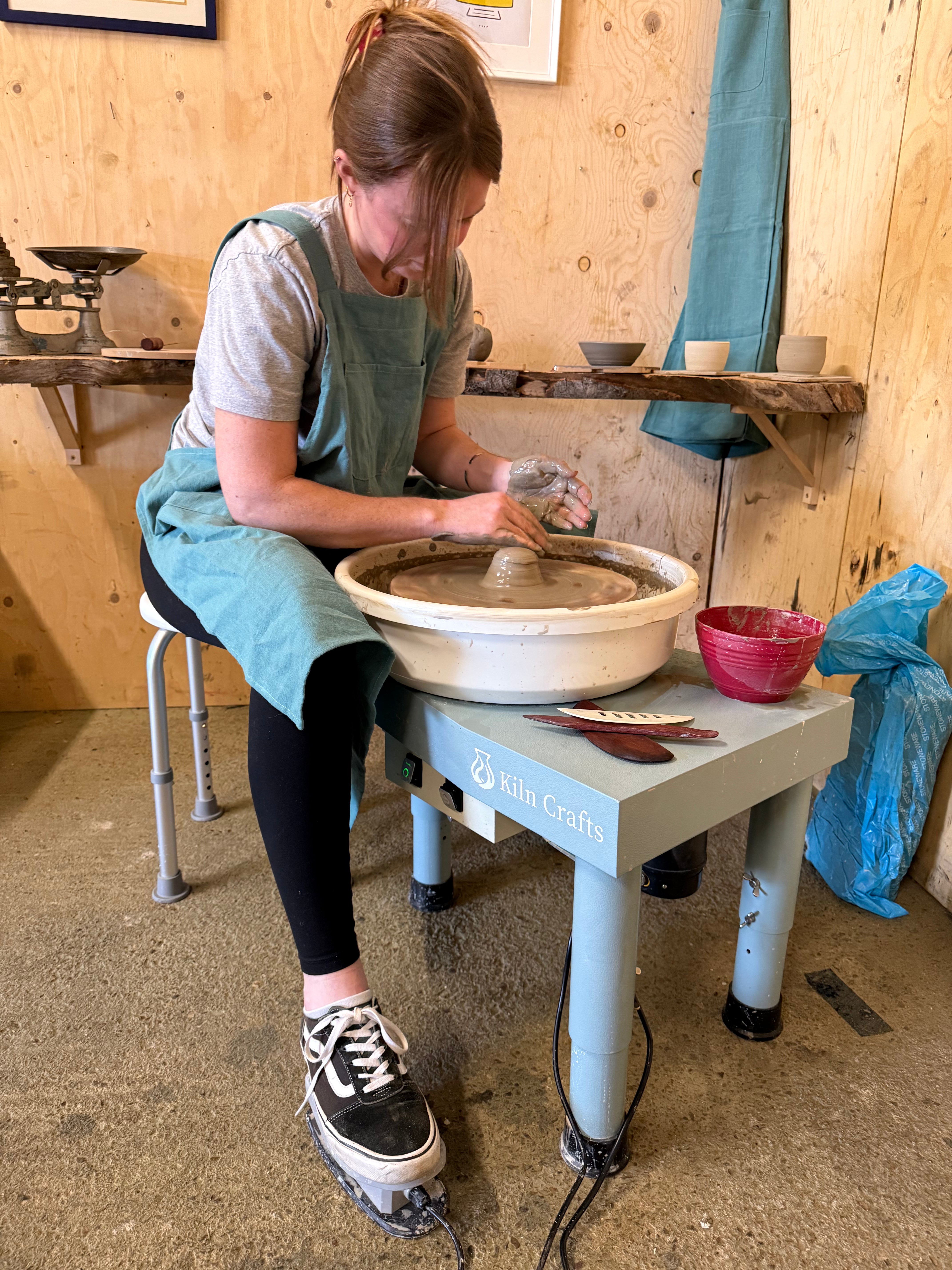 A female potter in a teal apron uses a Kiln Crafts pottery wheel to center clay. She is shown sitting on a height-adjustable stool, controlling the speed with a foot pedal while working in a bright, wood-panelled ceramic studio.