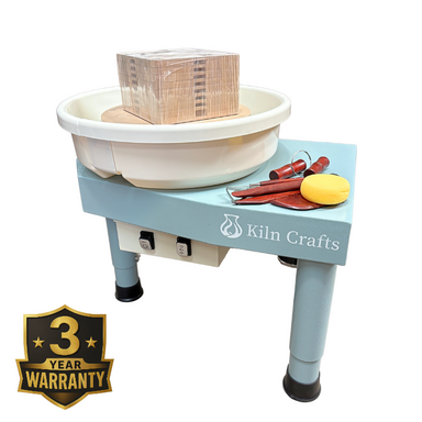 A Kiln Crafts pottery wheel with a light blue metal frame, white splash pan, and wooden bat. A set of wooden sculpting tools and a yellow sponge sit on the side table, and a 3-year warranty badge is featured in the corner.