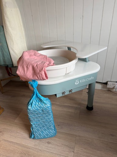 Kiln Crafts Thistle direct-drive pottery wheel with its shelf fitted, a pink craft apron and a bag of stoneware clay, set up at home